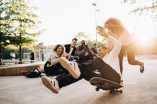 Lachende Freunde fotografieren Mann  der vom Skateboard fällt  während Frau ihn im Park schubst