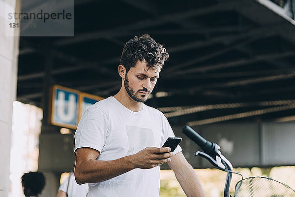 Junger Mann benutzt Smartphone  während er mit dem Fahrrad unter einer Brücke in der Stadt steht
