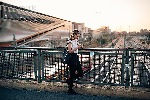 Junge Frau benutzt Mobiltelefon  während sie sich bei Sonnenuntergang an das Geländer einer Brücke in der Stadt lehnt