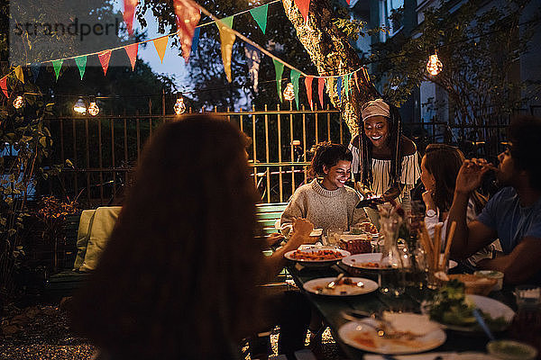 Lächelnde junge Frau zeigt Freundinnen bei Tisch während einer Dinnerparty ihr Handy