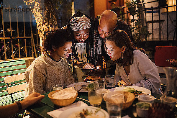 Glückliche junge Frau zeigt Freunden bei Tisch während der Dinnerparty ihr Smartphone