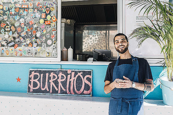 Porträt eines lächelnden jungen männlichen Verkäufers  der mit einem Smartphone gegen einen Speisewagen steht