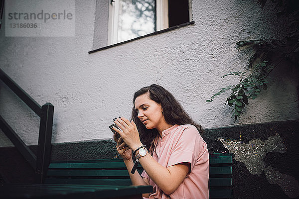 Junge Frau benutzt Mobiltelefon  während sie vor dem Haus auf einer Bank sitzt