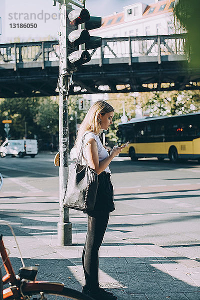 Seitenansicht einer jungen Frau  die ein Mobiltelefon benutzt  während sie auf dem Bürgersteig steht
