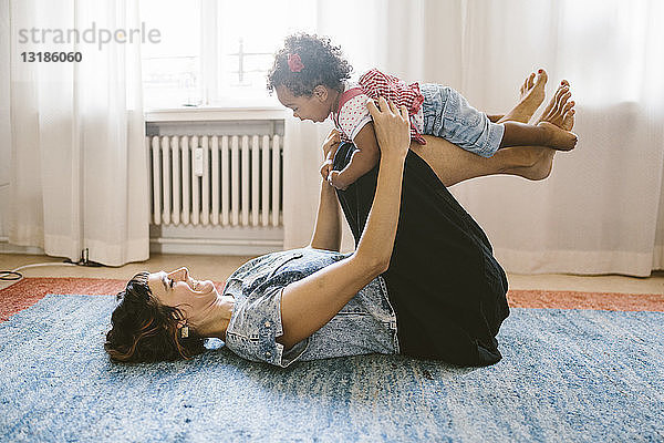 Verspielte Mutter  die ihre Tochter hebt  während sie zu Hause auf dem Teppich liegt