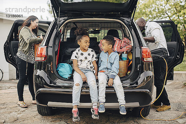 Lächelnde Geschwister in voller Länge auf dem Kofferraum eines Autos sitzend  während die Eltern im Vorgarten stehen