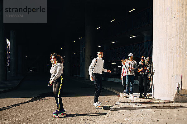 Freunde schauen Mann und Frau beim Skateboarden auf der Straße an einem sonnigen Tag zu