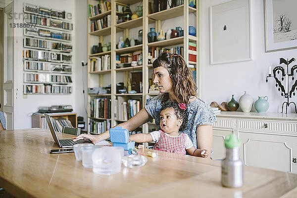 Frau benutzt Laptop  während sie mit ihrer Tochter am Esstisch im Haus sitzt