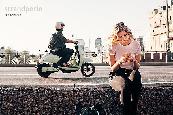 Lächelnde junge Frau  die ein Smartphone benutzt  während sie auf einer Stützmauer an einer Straße in der Stadt sitzt