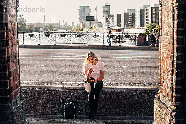Junge Frau benutzt Mobiltelefon  während sie auf einer Stützmauer an der Straße sitzt
