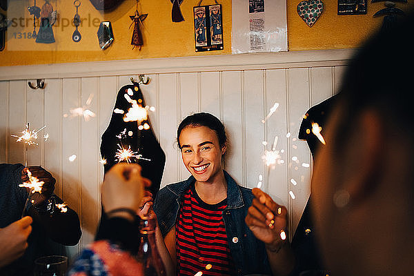 Lächelnde junge multiethnische Freunde halten brennende Wunderkerzen in der Hand  während sie während der Dinnerparty im Restaurant genießen