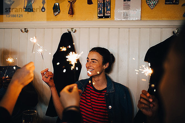 Lächelnde junge Frau hält brennende Wunderkerze in der Hand  während sie mit multiethnischen Freunden im Restaurant während einer Dinnerparty genießt