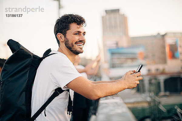 Seitenansicht eines lächelnden jungen Mannes  der wegschaut  während er sein Mobiltelefon an der Brücke hält