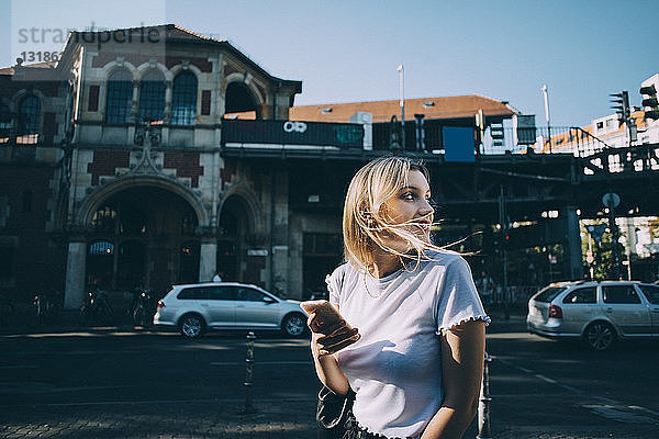 Junge Frau schaut über die Schulter  während sie ein Mobiltelefon auf der Straße in der Stadt hält