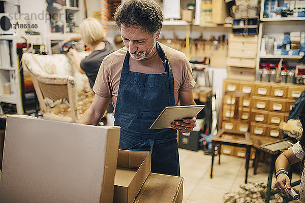 Männlicher Handwerker hält digitales Tablett in der Hand  während er in der Werkstatt einen Karton öffnet
