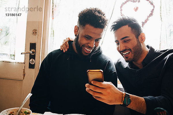 Lächelnde junge Männer teilen sich ein Smartphone  während sie während der Brunch-Party im Restaurant am Fenster sitzen
