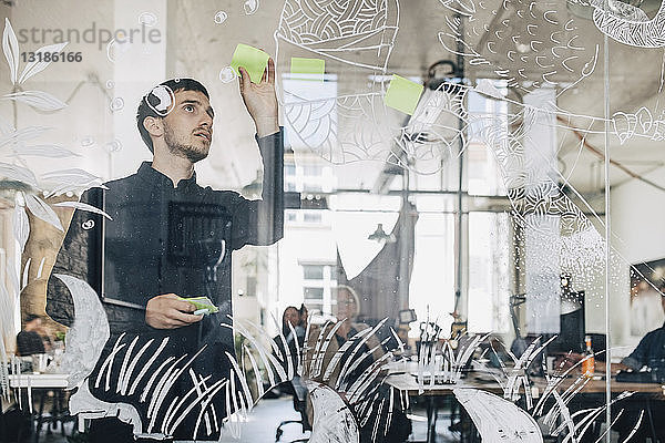 Junger kreativer Geschäftsmann klebt Klebezettel auf gemustertes Glas im Büro