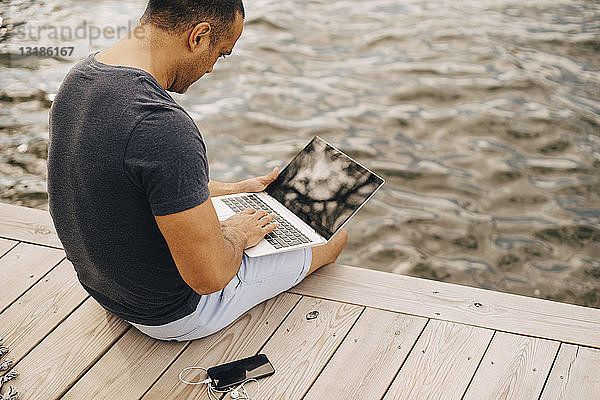 Erwachsener Mann sitzt mit Laptop auf dem Steg über dem See