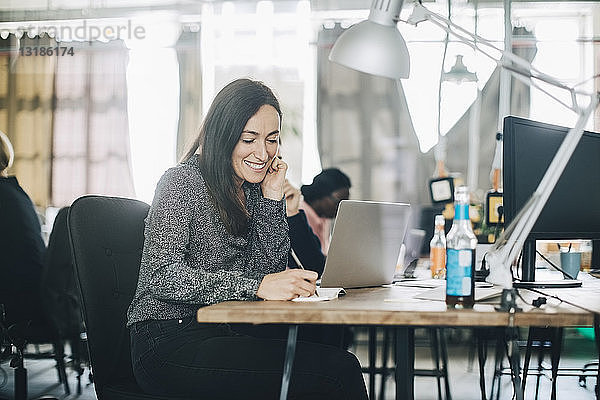 Kreative Geschäftsfrau lächelt beim Schreiben im Notizblock am Schreibtisch