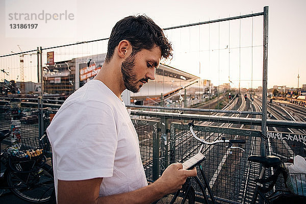 Seitenansicht eines jungen Mannes  der ein Mobiltelefon benutzt  während er auf einer Brücke über Eisenbahnschienen in der Stadt steht