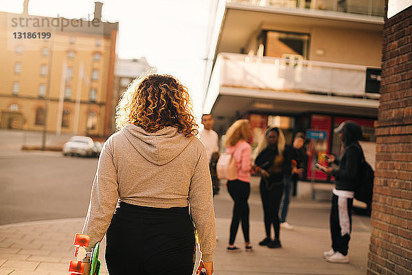 Rückansicht einer Frau mit Skateboard  die auf Freunde in der Stadt zugeht