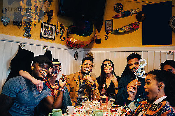 Porträt fröhlicher junger multiethnischer Freunde  die Requisiten halten  während sie während der Dinnerparty im Restaurant sitzen