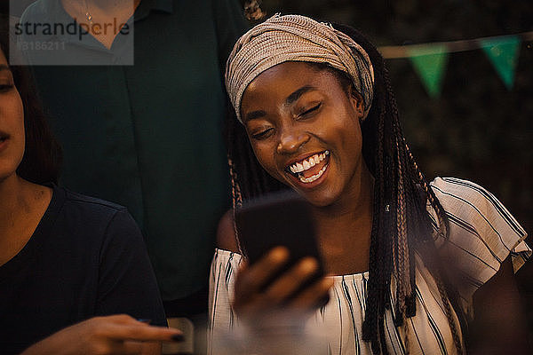 Junge Frau lacht  während sie während einer Dinnerparty mit Freunden im Hinterhof auf ihr Handy schaut