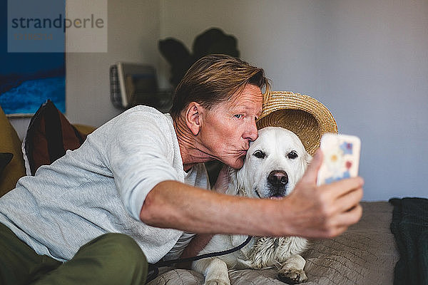 Älterer Mann küsst Hund  während er im Schlafzimmer zu Hause mit einem Smartphone Selbstgespräche führt