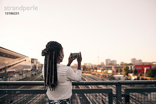Junge Frau fotografiert durch ein Mobiltelefon  während sie auf einer Brücke vor klarem Himmel steht