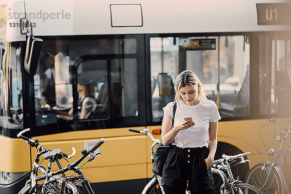 Junge Frau benutzt Smartphone  während sie in der Stadt an Fahrrädern gegen den Bus steht