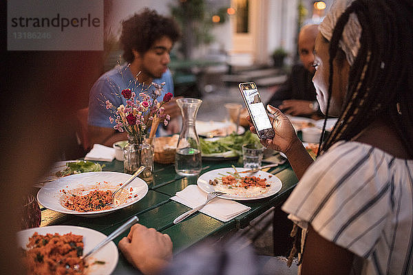 Junge Frau benutzt Mobiltelefon beim Abendessen mit Freunden während einer Gartenparty