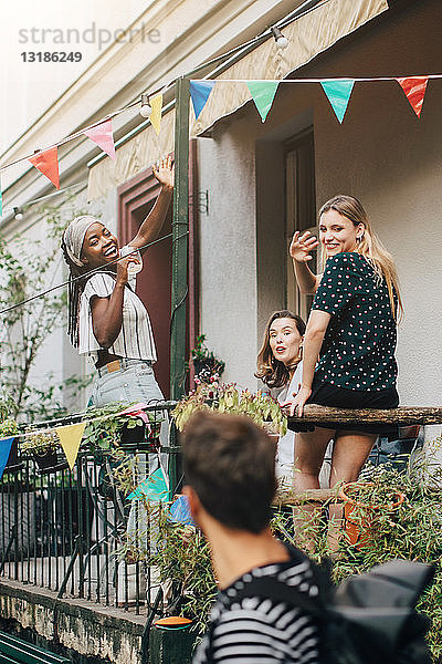 Glückliche junge Frauen winken einem männlichen Freund zu  während sie während einer Party auf dem Balkon stehen