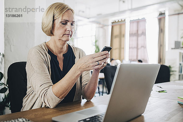 Reife Geschäftsfrau benutzt Mobiltelefon am Schreibtisch im Kreativbüro