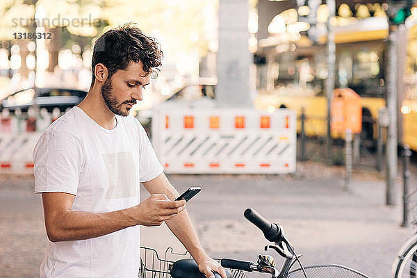Junger Mann benutzt Mobiltelefon  während er mit dem Fahrrad auf der Straße in der Stadt steht