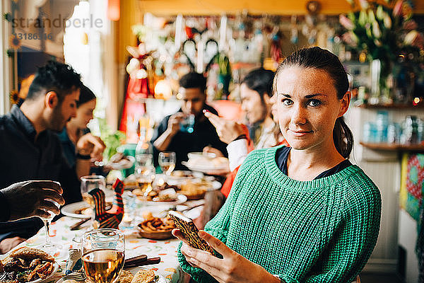 Porträt einer selbstbewussten jungen Frau  die ein Smartphone in der Hand hält  während sie mit Freunden am Tisch im Restaurant während des Abendessens sitzt