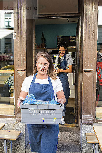 Lächelnde Besitzerinnen tragen Kisten gegen Restaurant