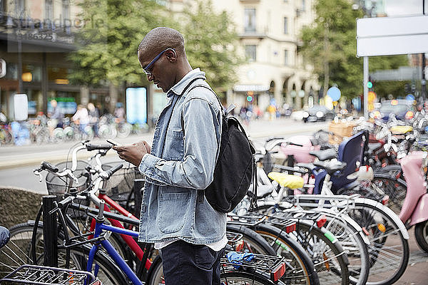 Seitenansicht eines mittelgroßen erwachsenen Geschäftsmannes  der ein Smartphone benutzt  während er in der Stadt an den Fahrrädern steht