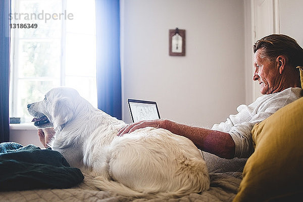 Älterer Mann benutzt Laptop  während er sich zu Hause neben dem Hund im Bett anlehnt