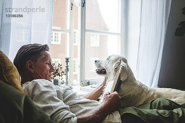 Älterer Mann liest Buch  während er sich zu Hause mit Hund im Bett entspannt