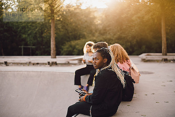 Nachdenkliche Teenagerin hält Handy in der Hand  während sie mit Freunden im Skateboard-Park sitzt