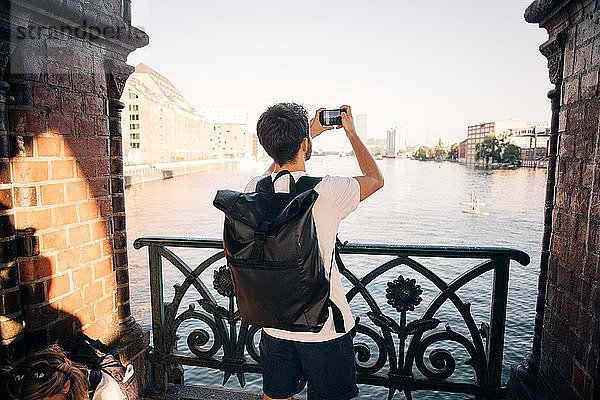 Rückansicht eines jungen Mannes  der mit einem Smartphone in der Stadt einen Fluss fotografiert