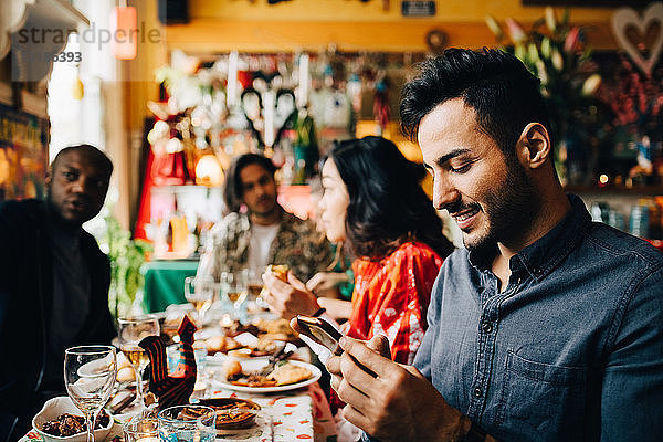 Lächelnder junger Mann sendet Textnachrichten auf einem Smartphone  während er während der Dinnerparty mit Freunden im Restaurant am Tisch sitzt