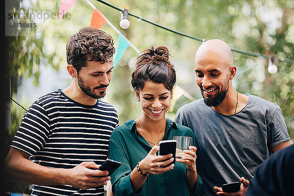 Glückliche junge Frau zeigt männlichen Freunden ihr Handy  während sie während einer Party auf dem Balkon steht