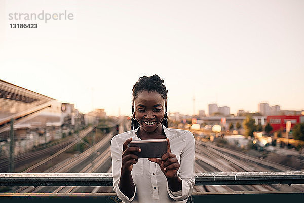 Lächelnde junge Frau fotografiert durch ein Mobiltelefon  während sie auf einer Brücke gegen den klaren Himmel in der Stadt steht