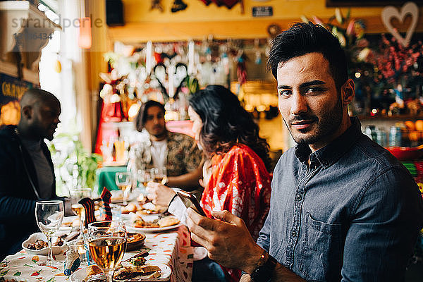Porträt eines selbstbewussten jungen Mannes  der ein Smartphone in der Hand hält  während er mit Freunden im Restaurant während des Dinner-Partes am Tisch sitzt