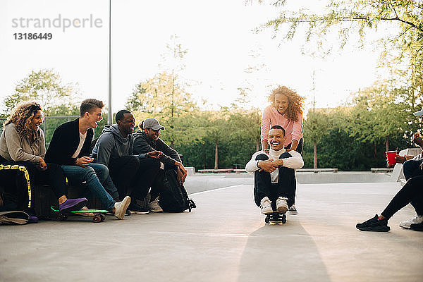 Freunde betrachten lächelnde Frau  die einen fröhlichen Mann schubst  der im Park auf einem Skateboard sitzt