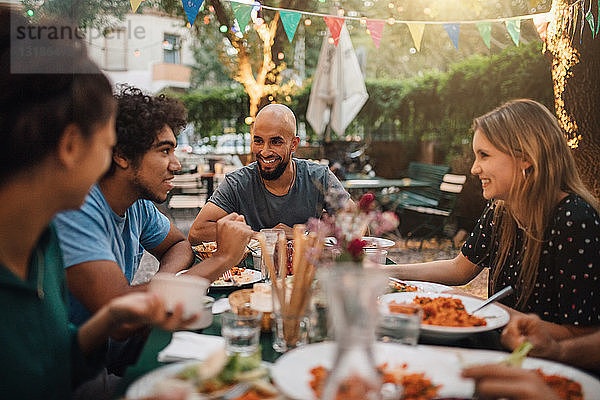 Lächelnder junger Mann und lächelnde Frauen lauschen dem Gespräch eines männlichen Freundes während einer Dinnerparty im Hinterhof