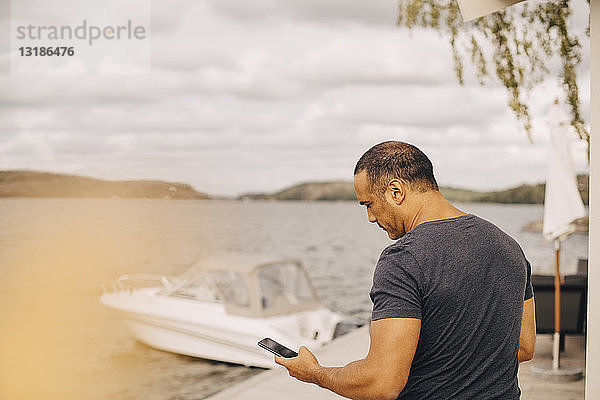 Ein erwachsener Mann benutzt ein Smartphone mit einer auf dem See vertäuten Yacht im Hintergrund