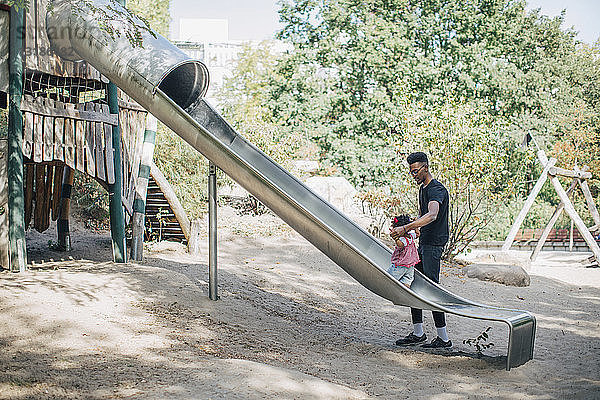 Vater hält Hände einer Tochter  die auf einer Rutsche auf einem Spielplatz läuft