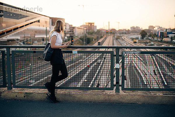 Seitenansicht einer jungen Frau  die bei Sonnenuntergang auf einer Brücke über Eisenbahnschienen in der Stadt steht